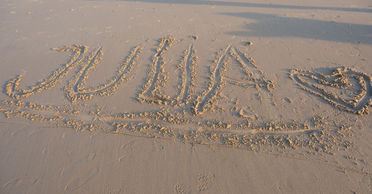 The name 'Julia' is artistically written in sand on a beach during sunset, with a heart symbol nearby.